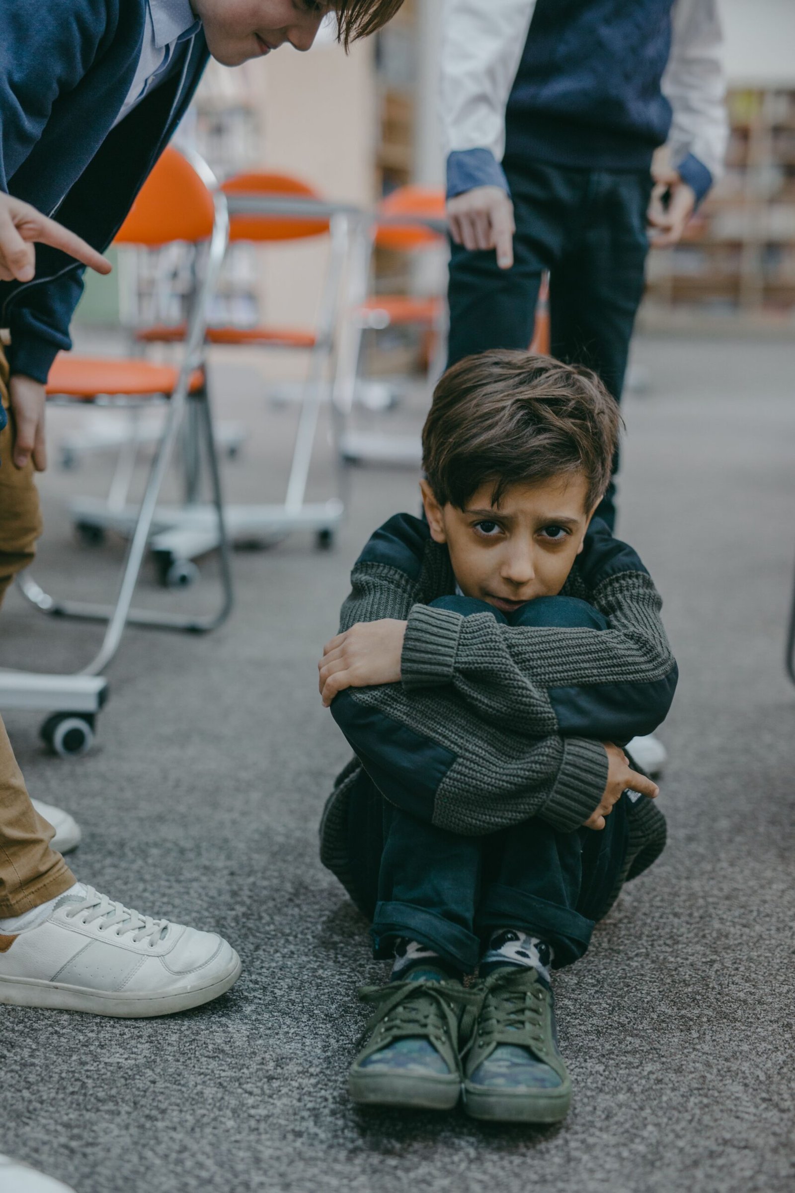 A sad boy sitting on the floor while children point at him in a classroom setting.